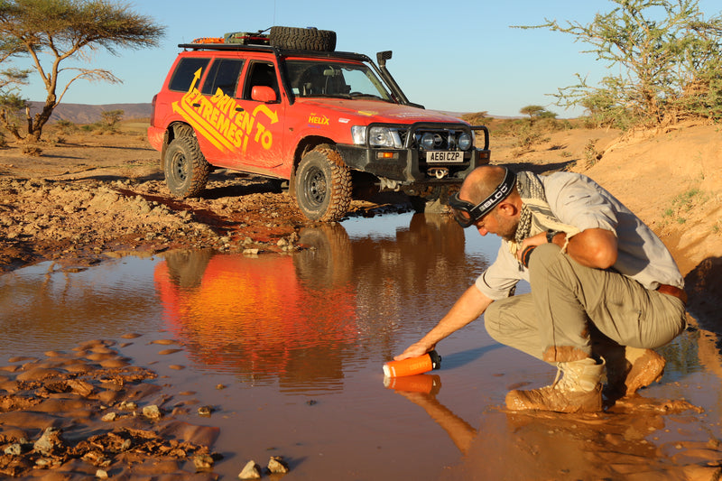 Laden Sie das Bild in Galerie -Viewer, Person refilling LifeSaver Liberty bottle from a muddle puddle by a red off-road vehicle in a desert setting
