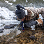 Person in winter clothing using a water filter straw by a stream.